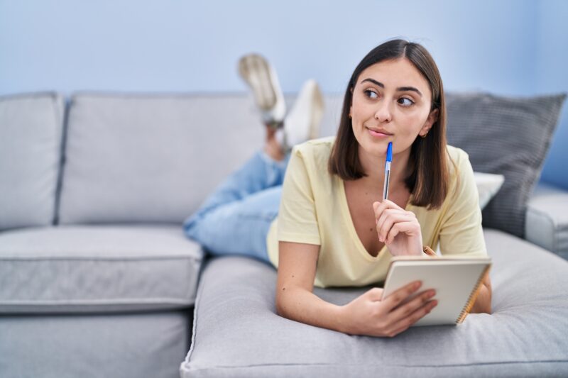 Young hispanic woman writing on notebook lying on sofa at home